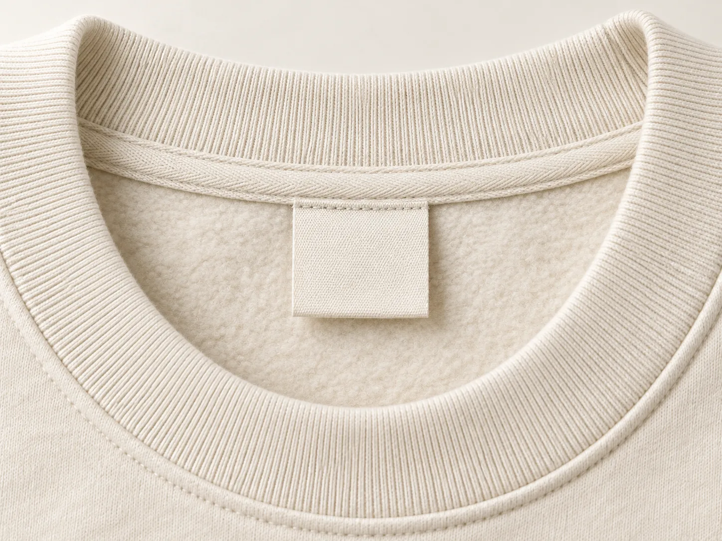 Close-up of a blank fabric label beneath the taped neckline of an ecru crewneck sweatshirt.