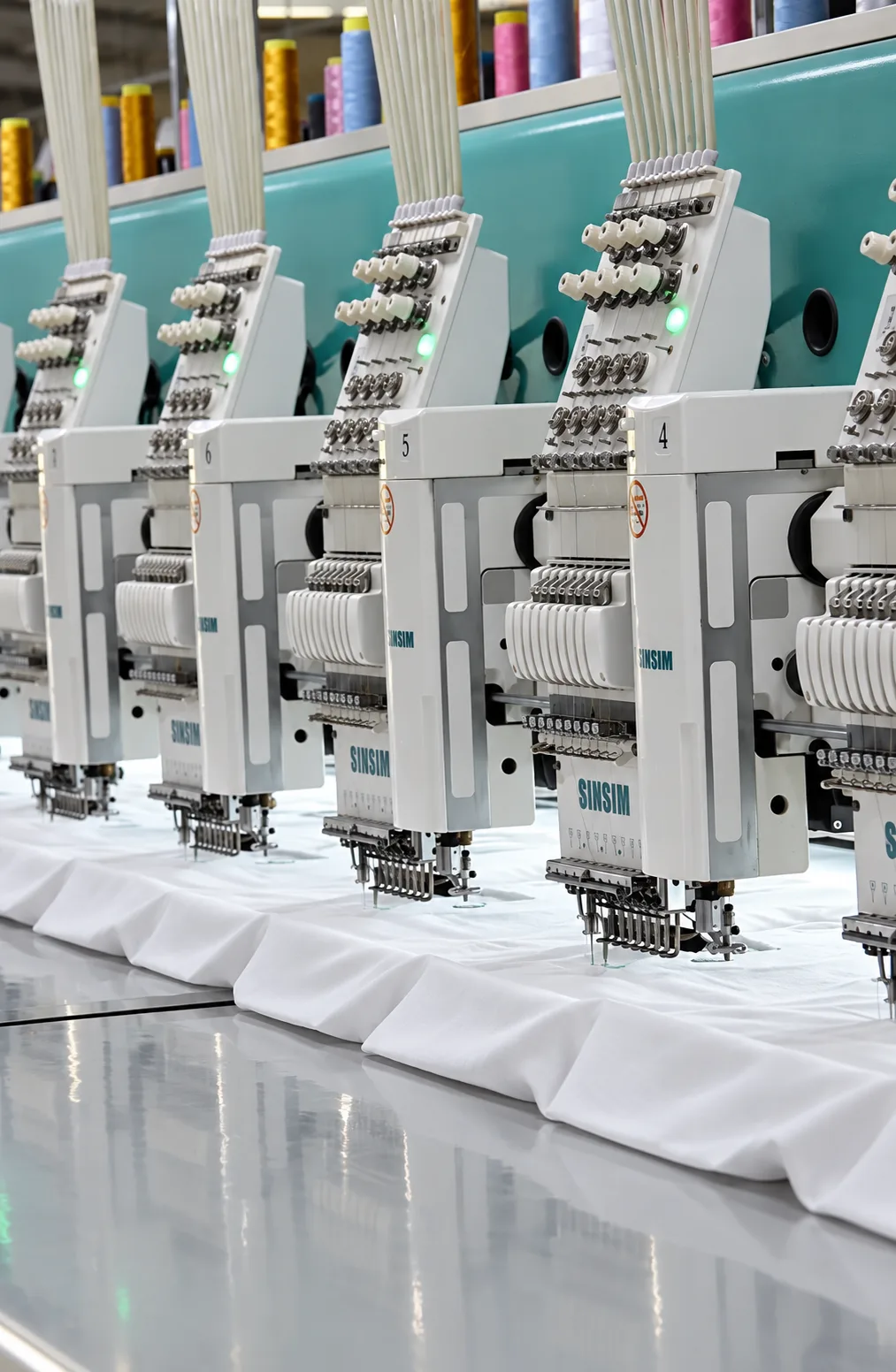 Embroidery heads and thread spools above white fabric held in frames.