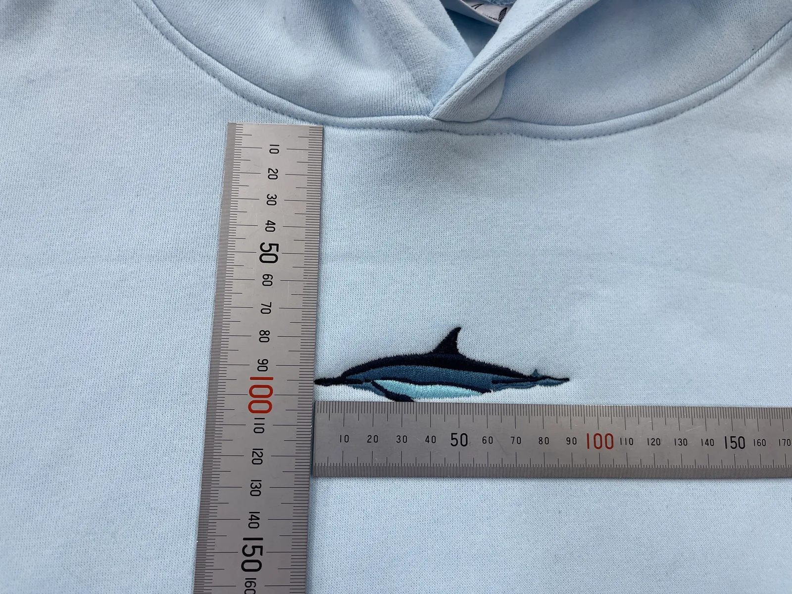 An embroidered fish motif on a pale blue hoodie beside two rulers.