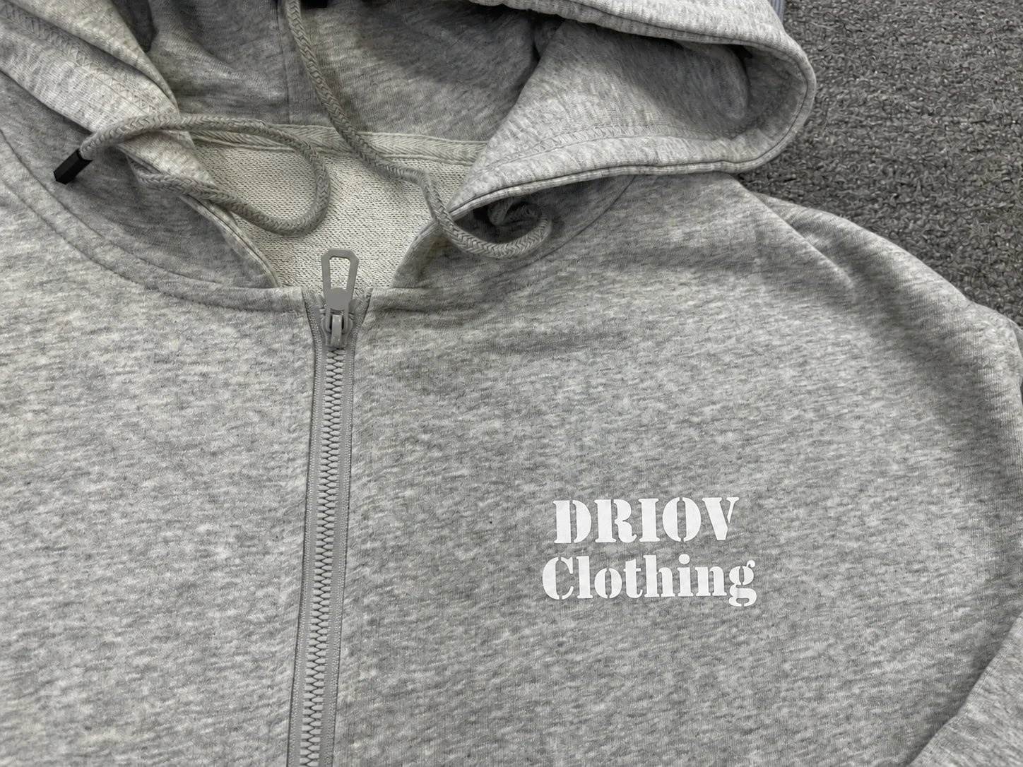White DRIOV Clothing lettering printed on the left chest of a grey zip hoodie.